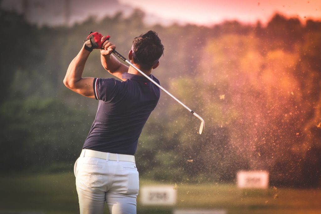 A man in a navy blue polo shirt and white pants is in the middle of a powerful golf swing, with a cloud of dirt and grass flying up behind him on a sunny golf course.