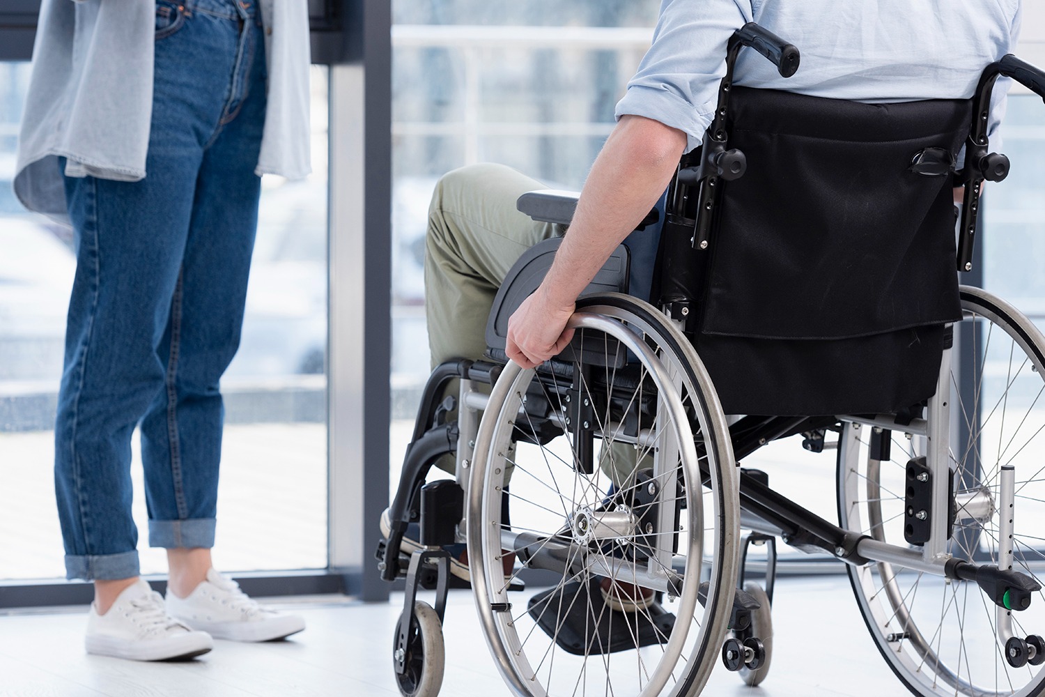 A man in a wheelchair is looking out a window. His hand is resting on the wheel of the wheelchair, and another person is standing nearby.
