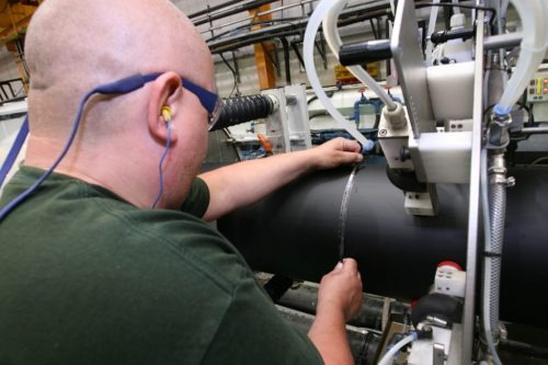 A bald man wearing safety glasses and earplugs is working on a large black pipe, with industrial machinery and hoses visible around him.