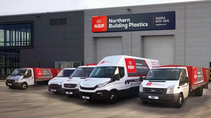 A fleet of white delivery vans from "Northern Building Plastics" is parked in front of a large grey warehouse building. The company logo is prominent on the building and the side of each van.