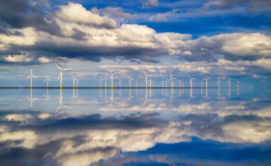 A row of offshore wind turbines standing on the horizon under a cloudy sky. The sky and turbines are reflected perfectly on the smooth water below.