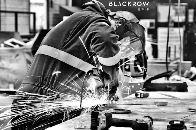 A black and white photo of a worker wearing a welder's helmet and safety suit, grinding metal, which creates a large shower of bright sparks. A power tool is on a table in the foreground.