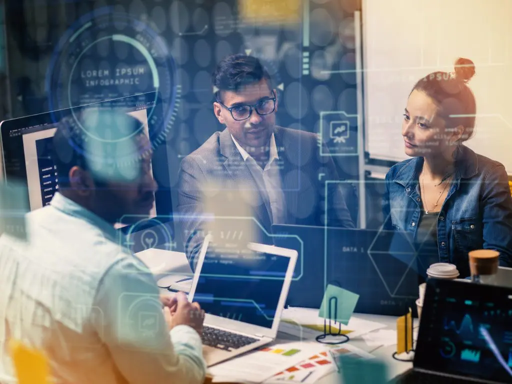 Three people are in an office meeting, appearing to discuss a project. Digital overlays with data and infographic elements are superimposed over the image, suggesting a technology or data-focused environment.