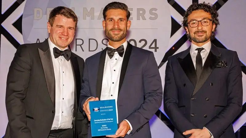 An image of three men smiling at the camera in front of an award background with the middle man holding an award.