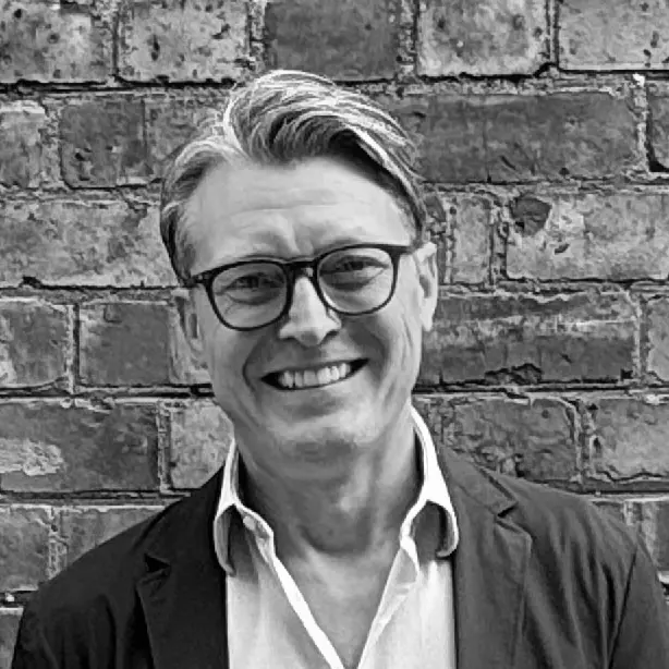 A black and white headshot image of a smiling man against a brick backdrop