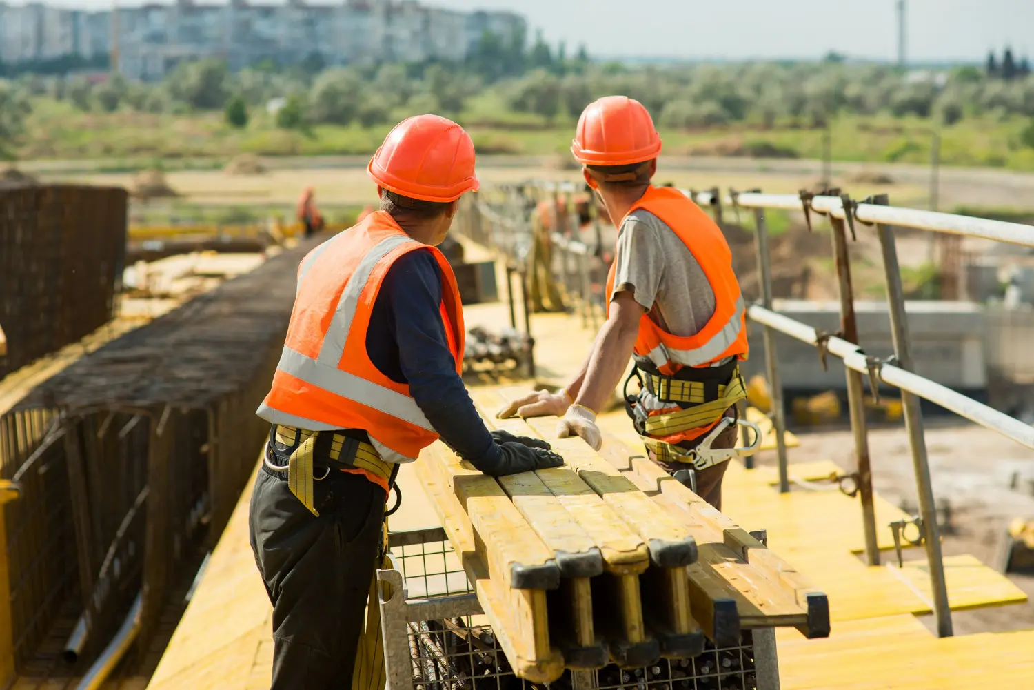 Two construction workers wearing safety vests and hard hats are handling a wooden plank on a building site.