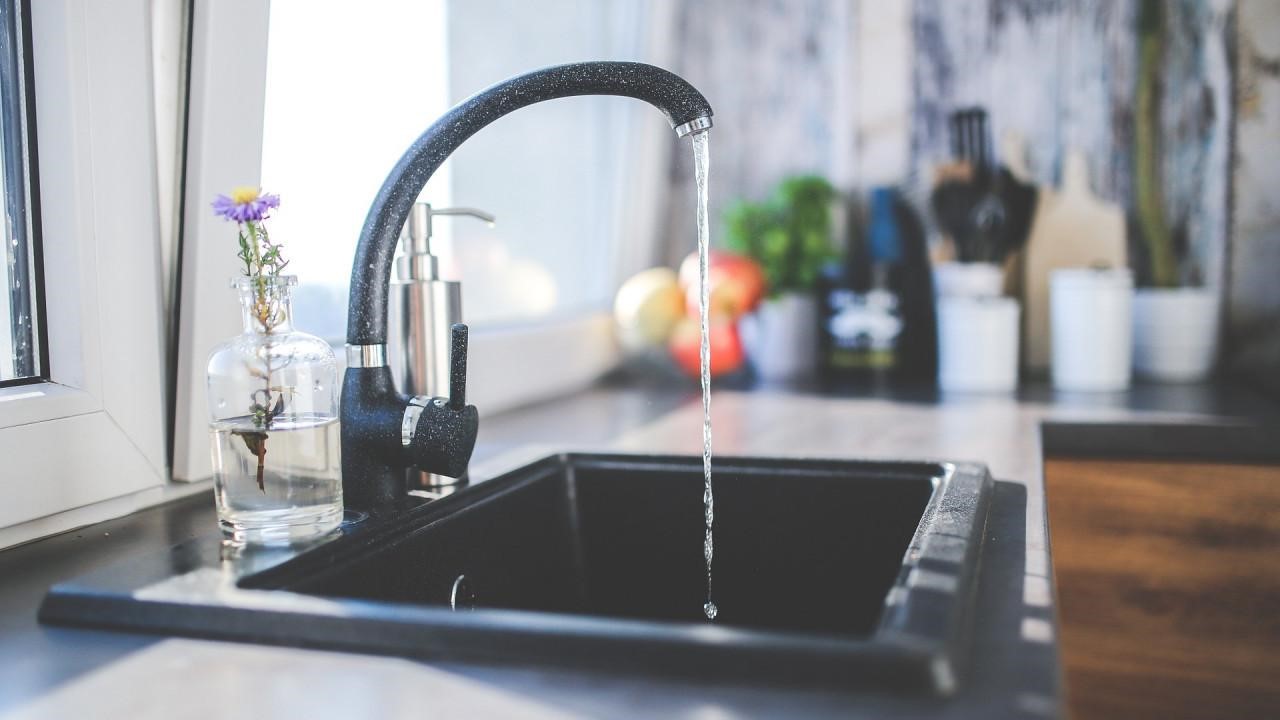 A close-up of a kitchen sink with a black, high-arc faucet. A stream of water flows from the faucet into the dark sink basin. A small vase with a flower sits on the windowsill to the left.