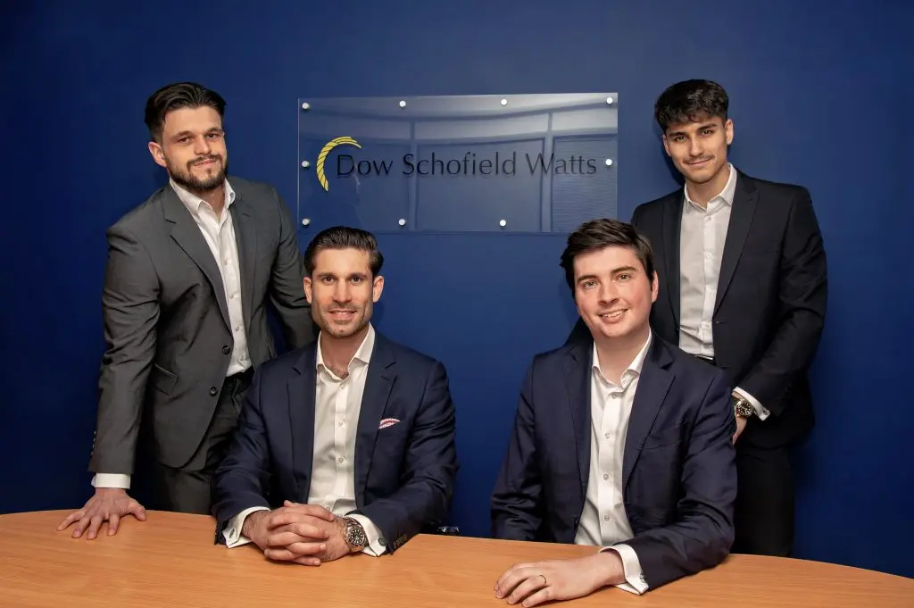 Two men standing behind two seated men smiling in front of the Dow Schofield Watts logo