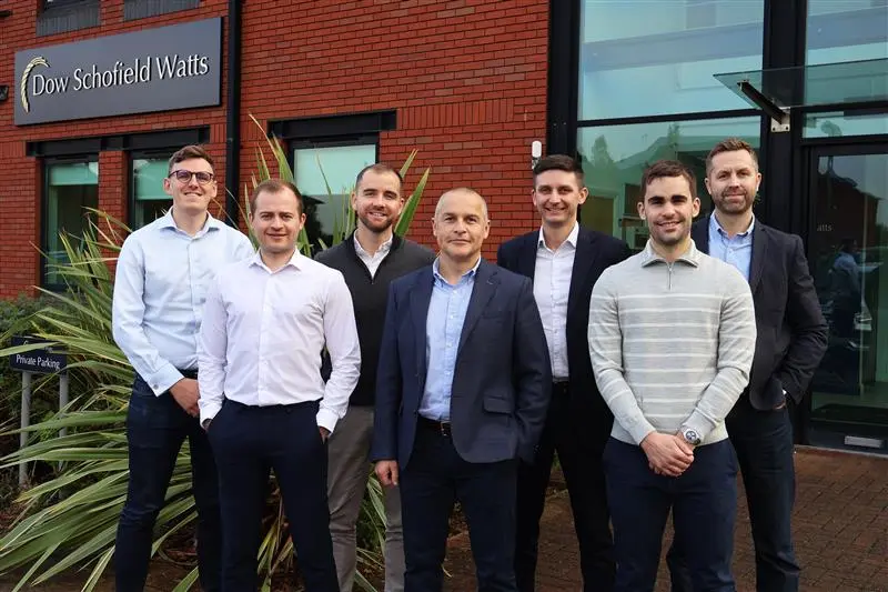 A team of seven male co-workers smiling and posing for a group photo in front of a brick office building. They appear happy and collaborative.