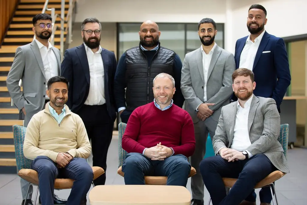 A diverse team of eight male co-workers smiling and posing for a group photo in a modern office. They appear happy and collaborative.