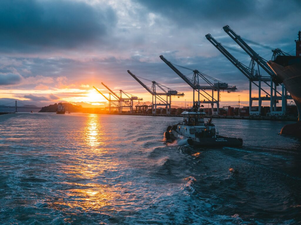 A tugboat moves across the water past large shipping cranes at sunset.