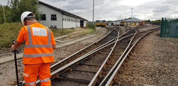 A person wearing an orange high-visibility vest and a white hard hat is working on a railway track with surveying equipment. The track splits into multiple directions, and a building and train are visible in the background.