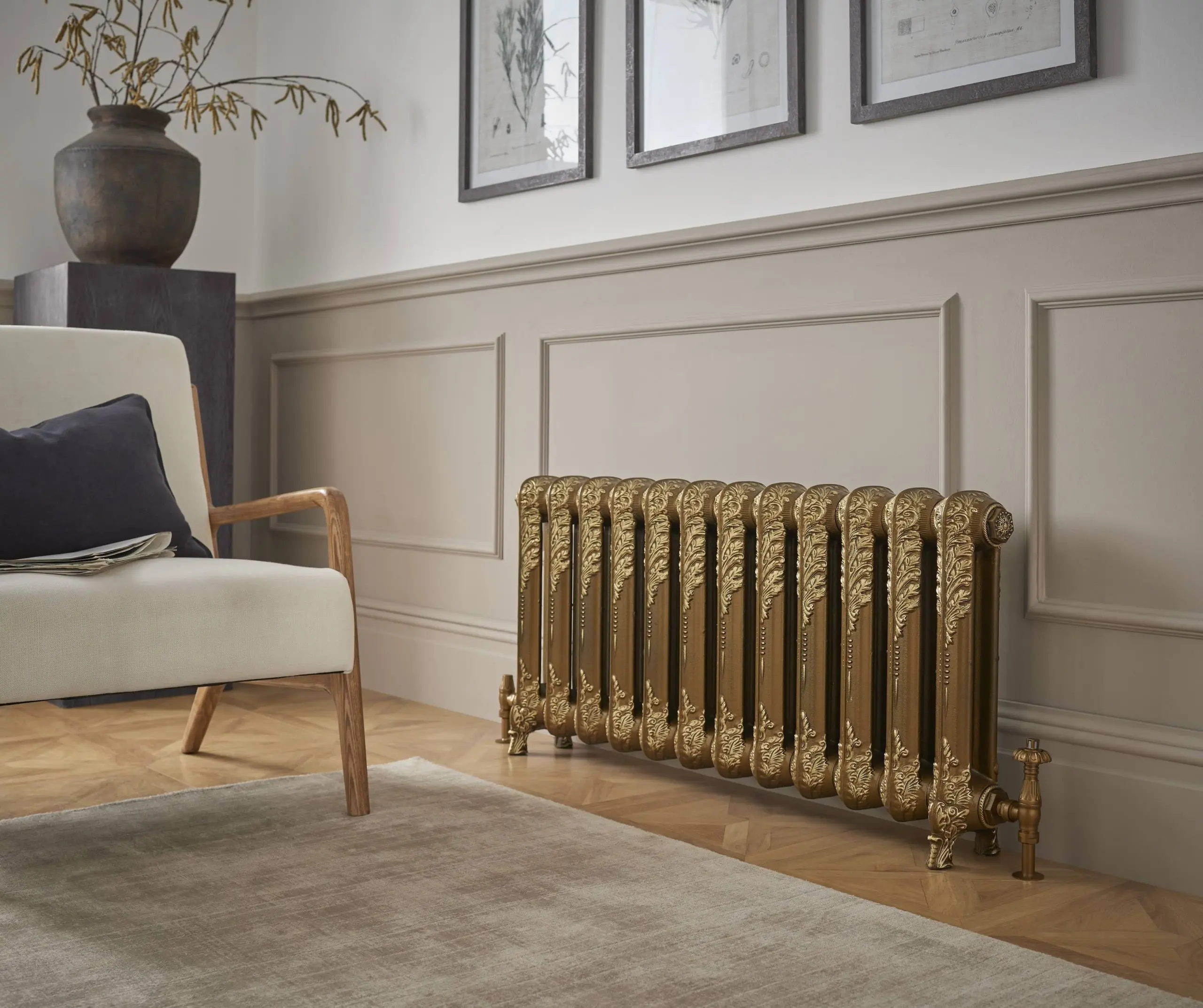A vintage, ornate, gold-coloured cast iron radiator sits on a light wooden floor. The radiator has detailed, decorative patterns and is positioned in front of a wainscot wall painted in a light tan or beige colour. In the background, there is a cream-colored armchair with a dark cushion, a large vase with branches, and three framed pictures hanging on the wall.