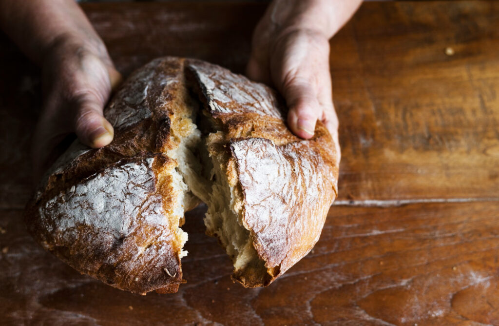 A close-up of a person's hands pulling apart a fresh loaf of rustic bread, revealing the soft, fluffy interior. The bread is on a dark wooden table.