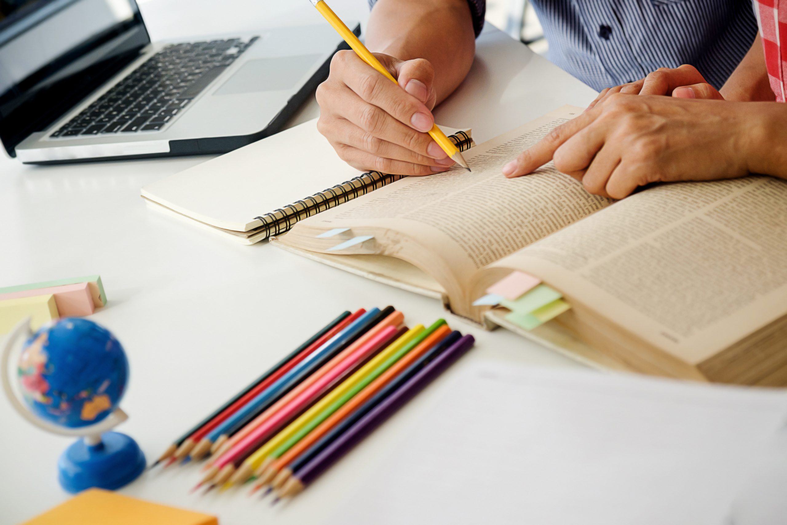 A close-up of two people's hands pointing at an open book with a yellow pencil. The desk also has a small globe, a handful of coloured pencils, and a laptop.