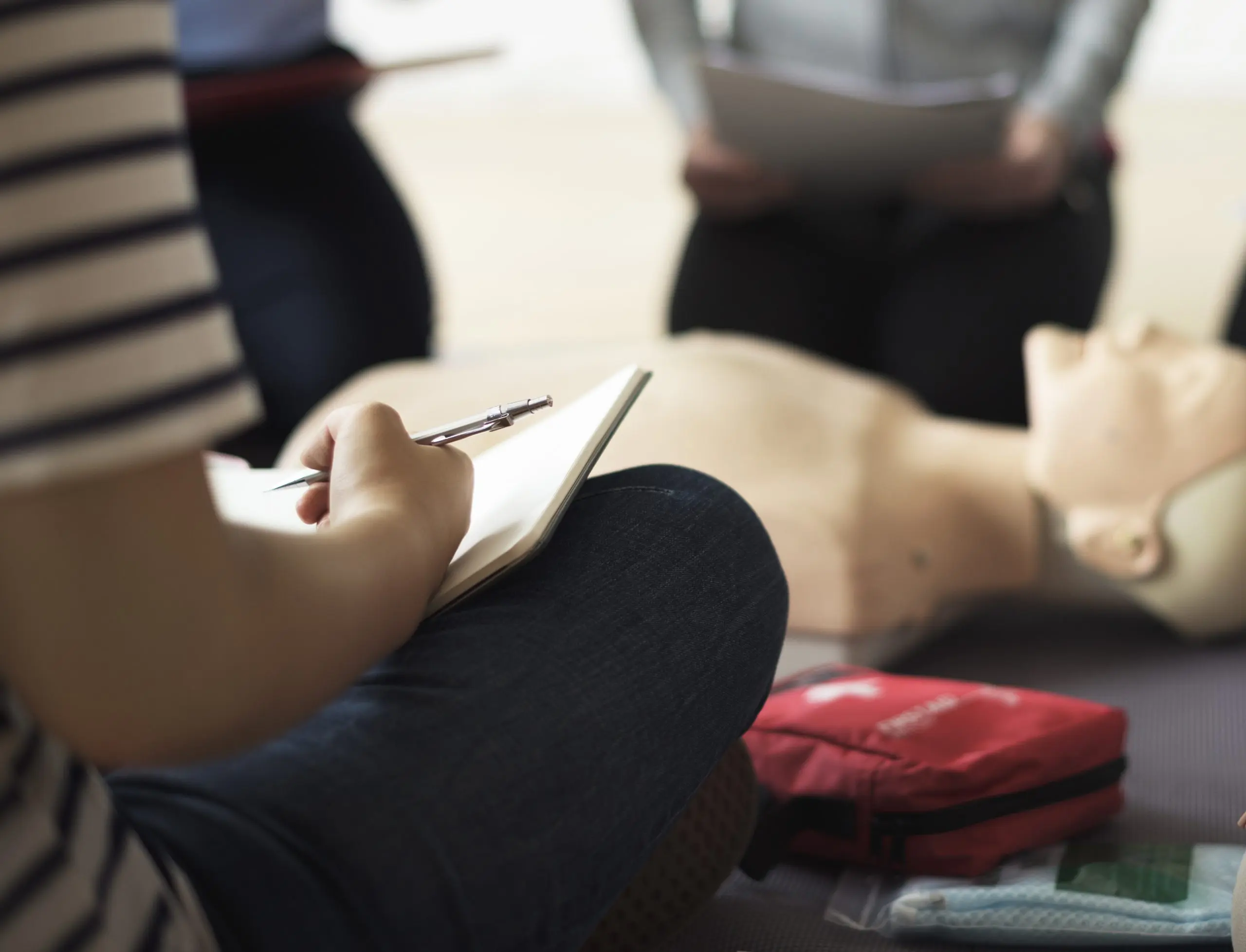 A close-up, high-angle shot of a person sitting and taking notes on a notepad during a first aid or CPR training course. In the background, a CPR mannequin lies on the ground next to a red first aid kit, while other people stand by.