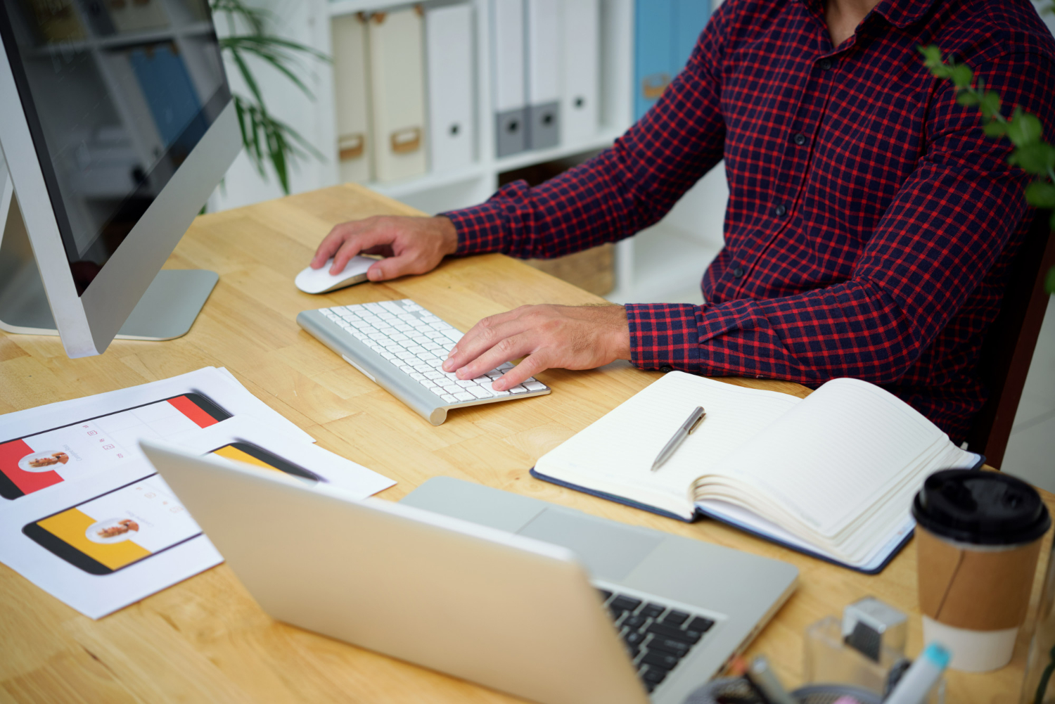 A man in a red plaid shirt is sitting at a wooden desk, working on a desktop computer. A laptop and a notebook are also on the desk.