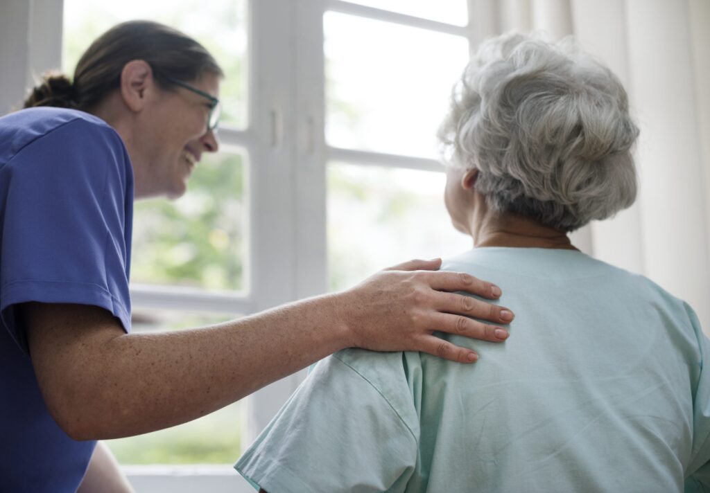 A nurse in a blue uniform stands and smiles at an elderly patient, placing her hand gently on the patient's shoulder. The patient, who has grey hair, is seen from the back, sitting and looking out a large window.