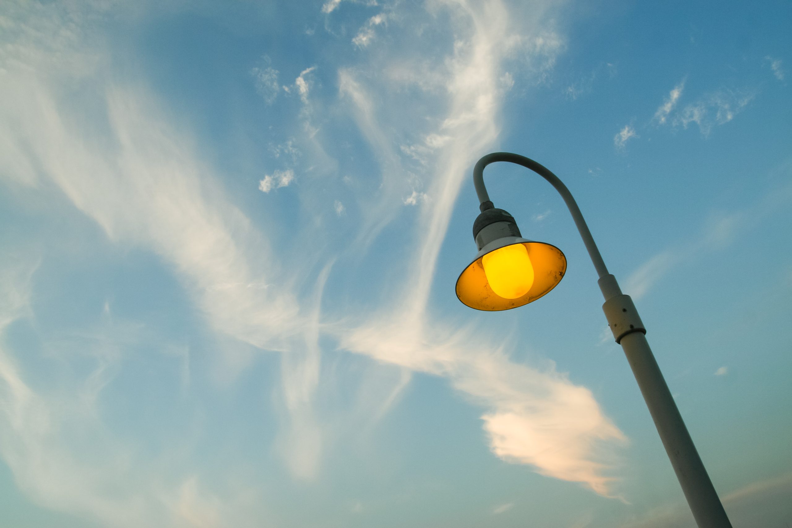 A lamppost with a yellow light is glowing against a bright blue sky with wispy white clouds.