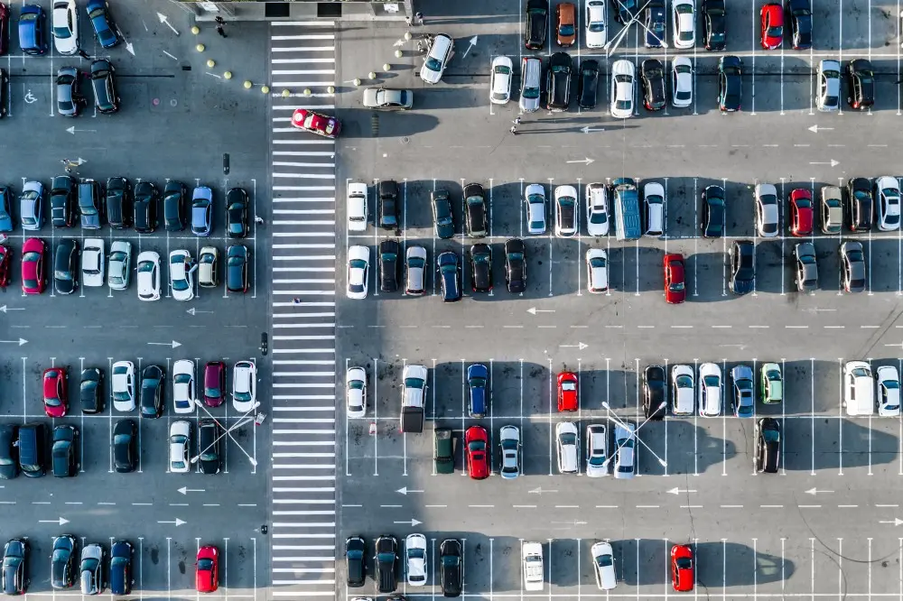An overhead shot of a large, busy parking lot. Cars of various colours and models are parked in rows, with a few empty spaces visible. There are also clear white lines indicating parking spaces, arrows showing traffic flow, and a zebra crossing.
