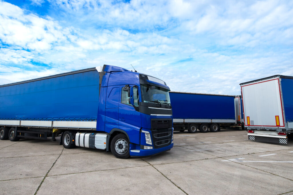 A fleet of large blue semi-trucks with matching blue cargo trailers parked in a lot under a bright, cloudy sky.
