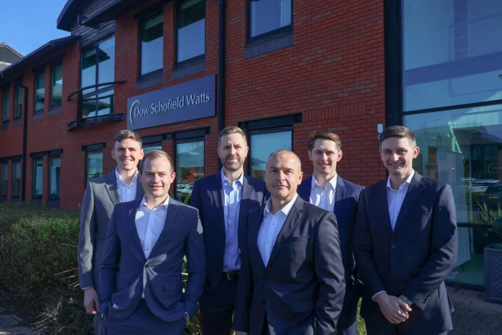 A group of seven professional men are standing and smiling in front of a red brick office building with a sign that reads "Dow Schofield Watts".