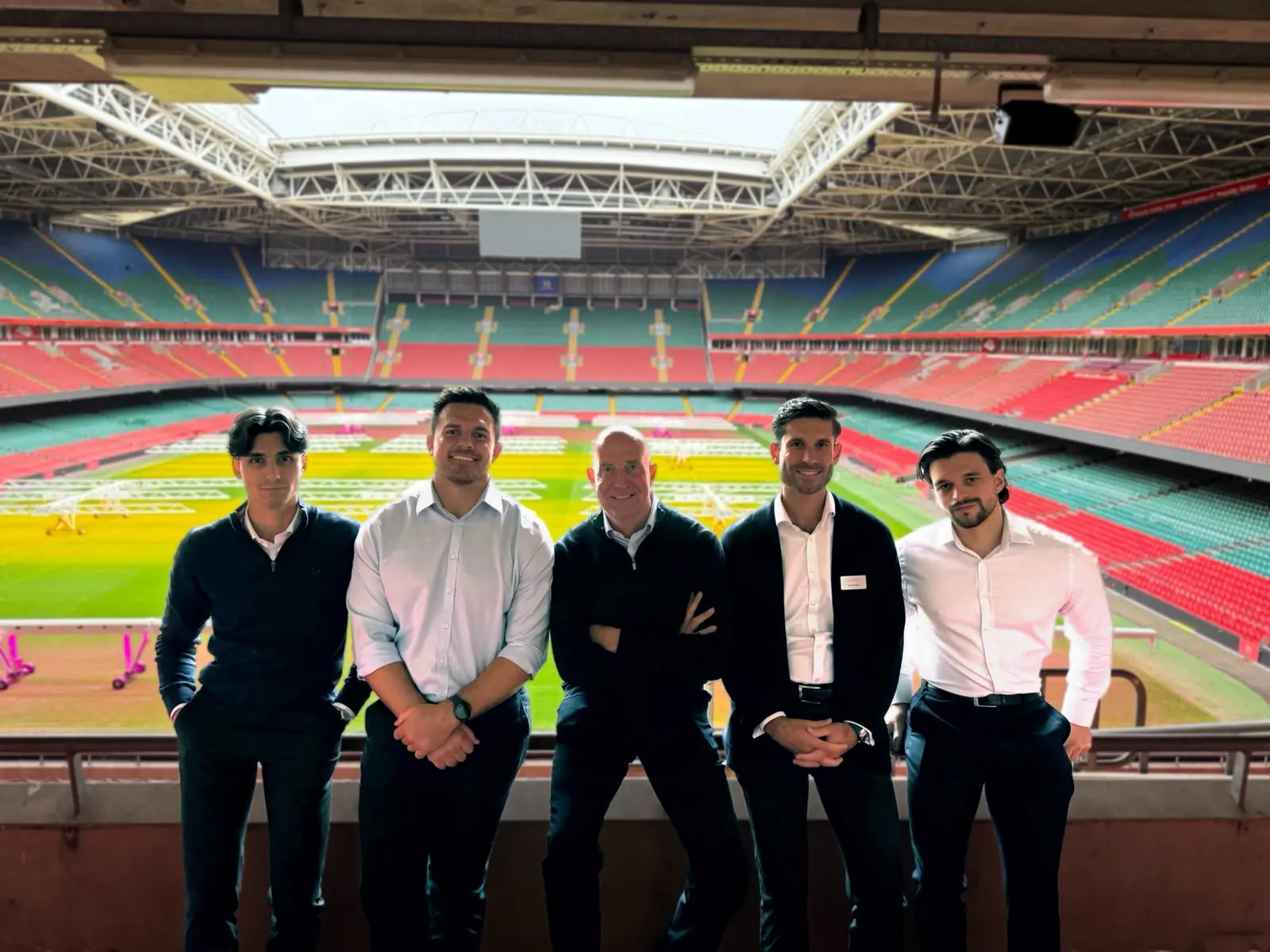 Five men in business wear smiling in front of a football stadium